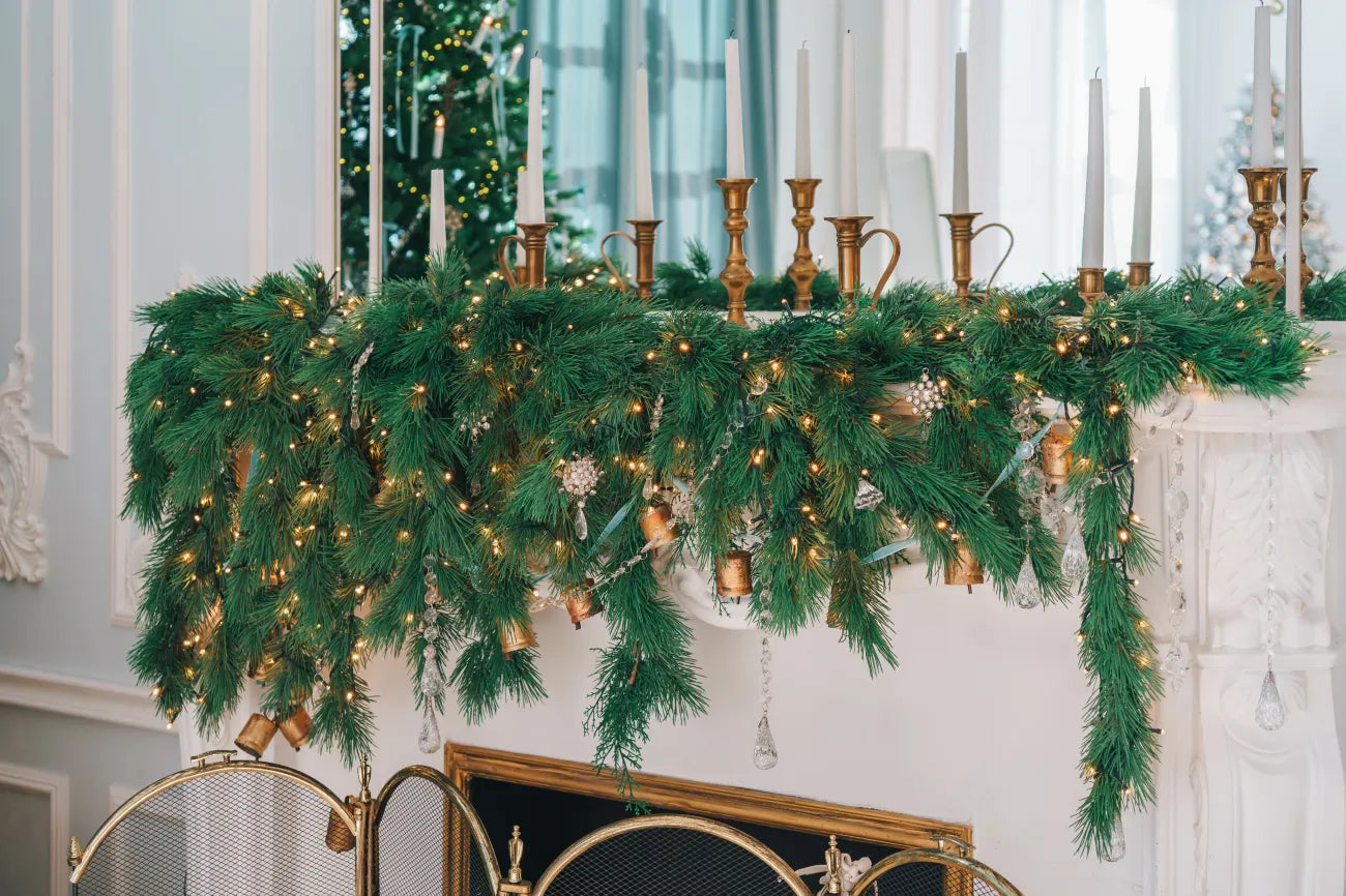 Beautifully decorated mantel is adorned with lush greenery garland with twinkling lights and golden candle holders with candles.