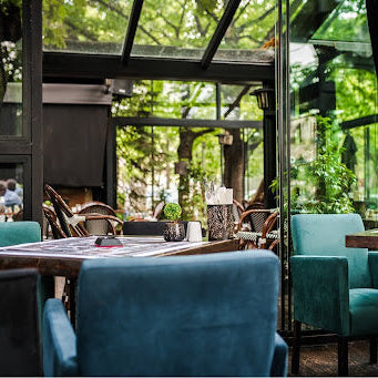 Modern restaurant interior with teal chairs, dark wood tables, and floor-to-ceiling windows overlooking greenery.