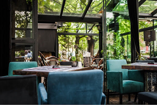 Modern restaurant interior with teal chairs, dark wood tables, and floor-to-ceiling windows overlooking greenery.